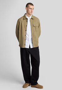 Giacca khaki con tasca frontale, indossata sopra una camicia bianca, abbinata a pantaloni neri e mocassini marrone chiaro su uno sfondo neutro.