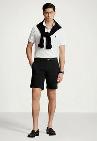 Νεαρός άνδρας που φοράει λευκό πόλο μπλουζάκι, μαύρες βερμούδες, μαύρο πουλόβερ ριγμένο στους ώμους, μαύρη ζώνη, ρολόι και μοκασίνια.