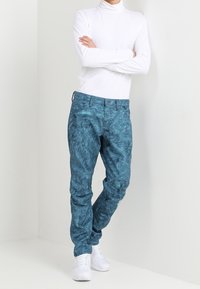 Turquoise jeans with textured fabric and a slim fit, paired with a fitted white turtleneck and white sneakers. Simple design with no visible patterns.