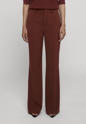 Persona che indossa pantaloni a vita alta e gamba larga color ruggine, abbinati a décolleté nere con punta a punta, in piedi davanti a uno sfondo chiaro e semplice.