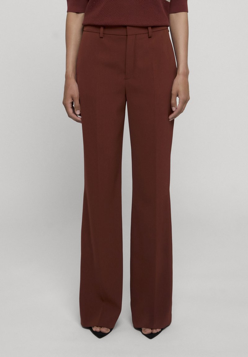 Personne portant un pantalon taille haute à jambes larges couleur rouille, associé à des talons noirs pointus, debout devant un fond clair uni.
