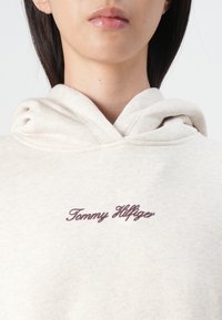 Hoodie boje kreme napravljen od mekog materijala, s prednjim džepom i ušivenim logotipom u tamnosmeđoj boji. Kapuljača ima opušteni kroj i vidljive šavove.