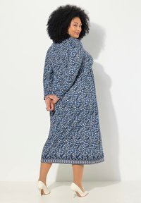 Niebieska sukienka w kwiatowy wzór z długimi rękawami, luźny krój oraz białymi akcentami u dołu, zestawiona z beżowymi szpilkami.