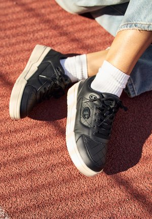 Osoba nosząca czarne skórzane sneakersy z białymi podeszwami i białe skarpetki, siedząca na czerwonym, teksturowanym podłożu, ubrana w jasnoniebieskie dżinsy.