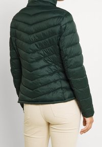 Vihreä tikkikangas puhvijakku, jossa on korkea kaulus, ja jossa on viivoituskuvio ja joustavat hihansuut. Yhdistetty vaaleanbeigeihin housuihin.