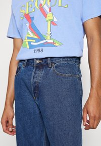 Světle modré grafické tričko s textem "SEOUL" a multikolorovým designem, v kombinaci s tmavomodrými džíny z denimového materiálu a oranžovými švy.