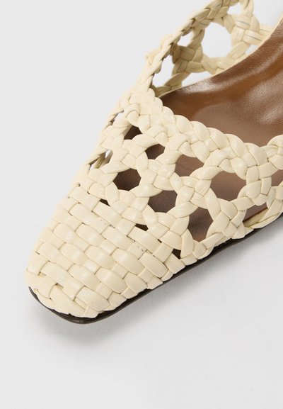 Zapato de cuero tejido en color beige con diseño de punta afilada, lados abiertos y suela plana. Presenta un patrón texturizado en forma de cruz.