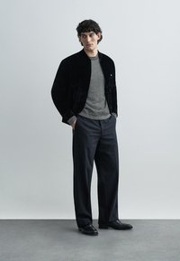 Crna baršunasta bomber jakna preko sive prugaste majice, uparena s širokim tamnim hlačama i crnim cipelama, na jednostavnoj pozadini.