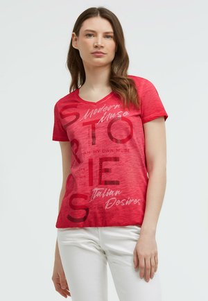 Junge Frau mit langen braunen Haaren, die ein rotes T-Shirt mit Grafik und Text sowie weiße Hosen vor einem schlichten hellen Hintergrund trägt.