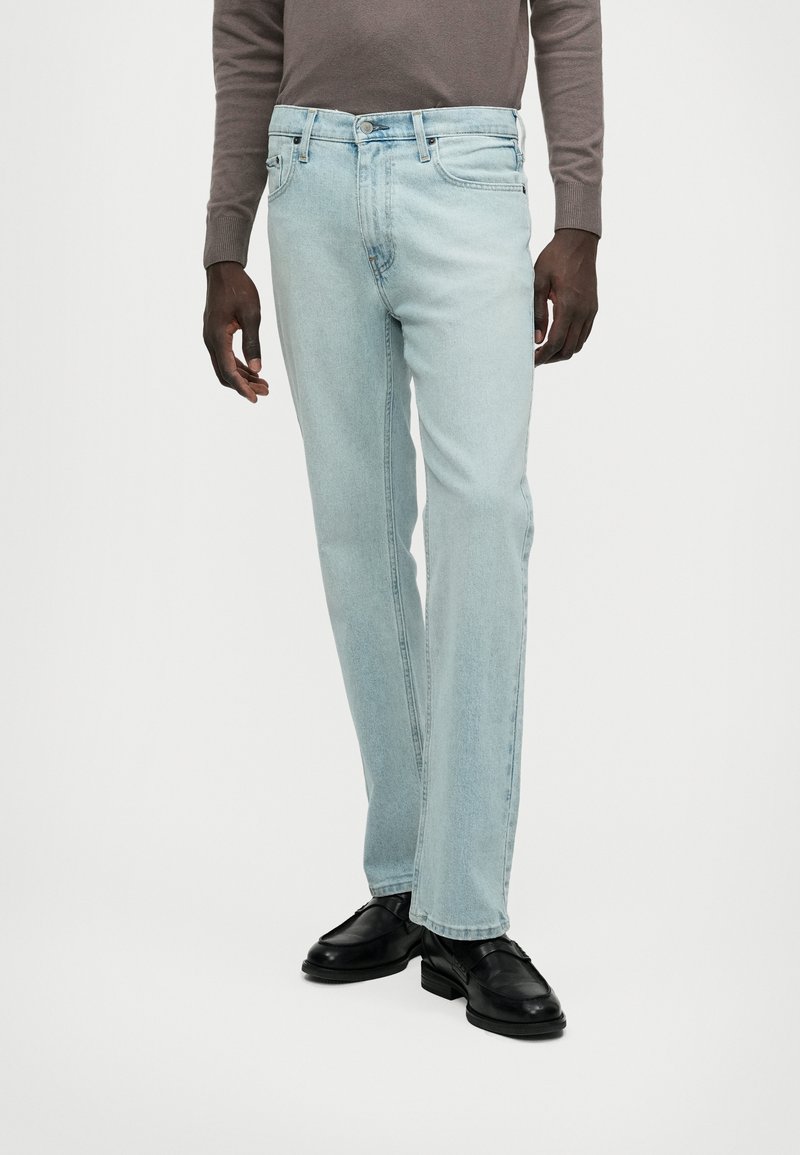 Ανοιχτού μπλε τζιν παντελόνι με ίσια γραμμή, φτιαγμένο από ντένιμ, που διαθέτει πέντε τσέπες και φερμουάρ με κουμπί. Φοριέται με μαύρα παπούτσια.