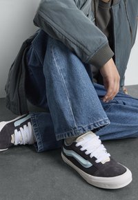 Svart og lyseblå sneakers med hvite snører, med en overdel i semsket lær og skinn, satt mot blå denimjeans og en grå bomberjakke.