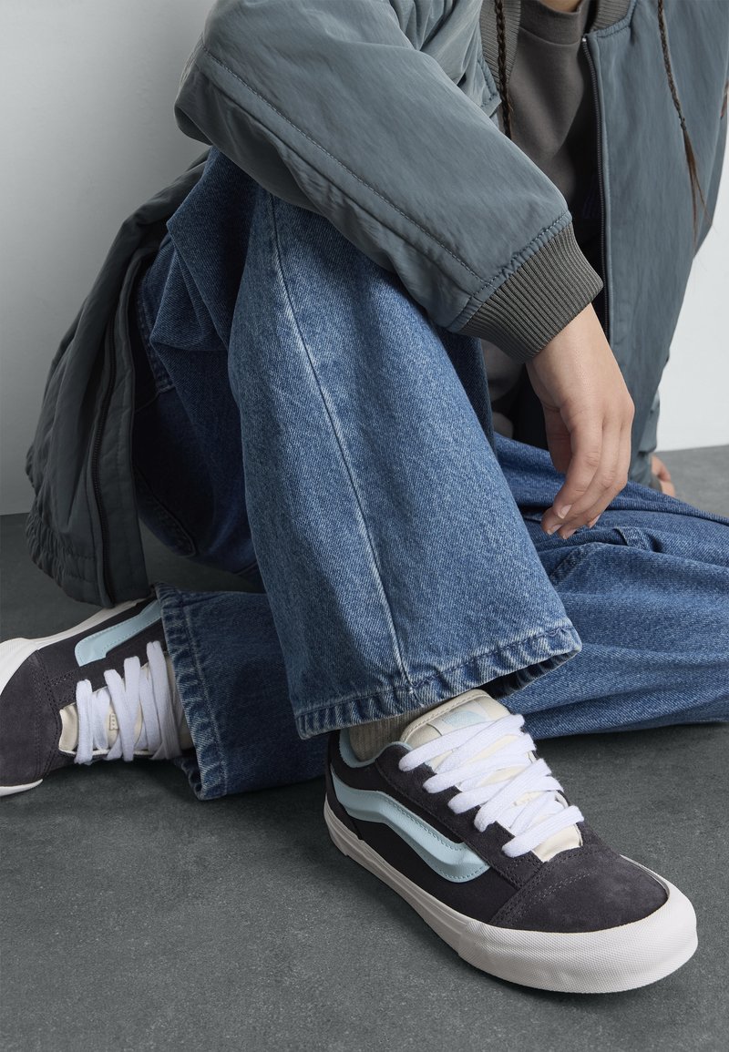 Svart og lyseblå sneakers med hvite snører, med en overdel i semsket lær og skinn, satt mot blå denimjeans og en grå bomberjakke.