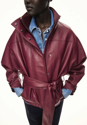 Persona que lleva una chaqueta de cuero burdeos con cinturón sobre una camisa vaquera azul con botones, con las manos en los bolsillos de la chaqueta.