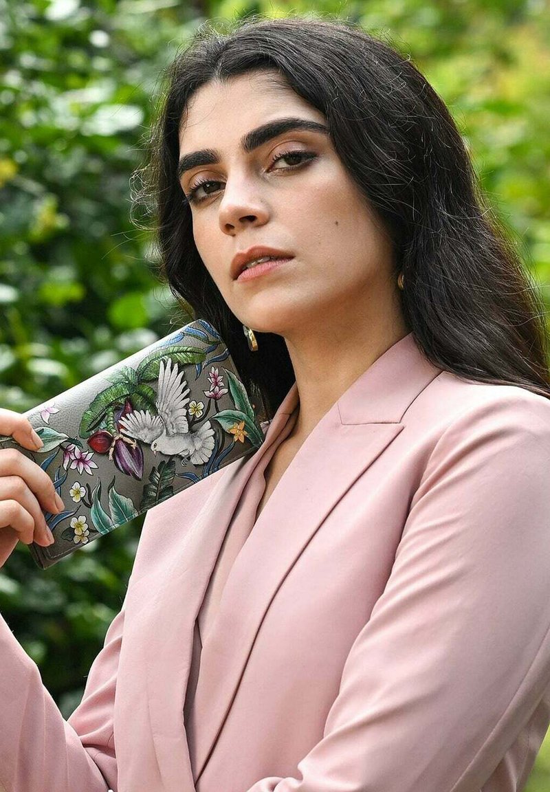 Kleurvolle leren portemonnee met een gedetailleerd bloemen- en vogelontwerp, vastgehouden door een persoon in een lichtroze blazer. Soepele textuur, rechthoekige vorm.