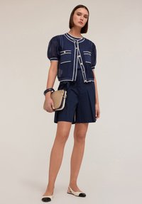 Navyblauwe jas met pofmouwen en witte afwerking, gecombineerd met navy shorts. Model houdt een beige tas met gouden hardware vast en draagt crèmekleurige en zwarte ballerina's.