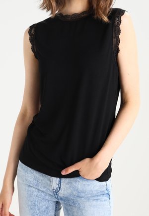 Vrouw draagt een mouwloos zwart topje met kanten rand en lichtblauwe spijkerbroek, hand in de zak, tegen een witte achtergrond.