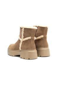 Botas de ante marrón hasta el tobillo con ribete de piel sintética de borreguito, suela de plataforma plana y costuras decorativas. Presentan un diseño de cruz asimétrico.