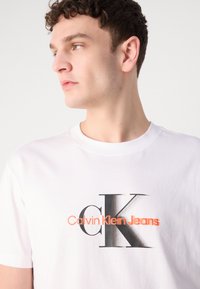 Hvit bomulls t-skjorte med et stort grafisk trykk i svart og oransje som leser "Calvin Klein Jeans" med et fremtredende "CK"-design.