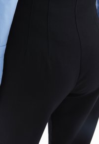 Černé elastické kalhoty z hladké tkaniny, s plochými předními kapsami a vysokým pasem, doplněné světle modrou košilí.