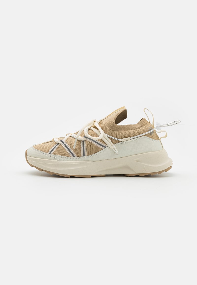 Zapatilla deportiva beige con parte superior de malla, acentos en crema claro y suela de goma texturizada. Presenta cordones elásticos y una lengüeta en la parte trasera.