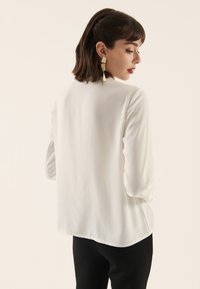 White long-sleeve blouse with a smooth texture, round neckline, and slightly curved hem, paired with black pants.