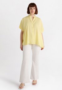 Blusa gialla a maniche corte con colletto a punta e bottoni frontali, abbinata a pantaloni bianchi svasati e tacchi bianchi con cinturini.