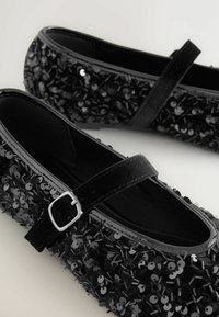 Las bailarinas negras presentan un upper con lentejuelas y un acabado texturizado, una correa con hebilla plateada y un interior liso y oscuro.