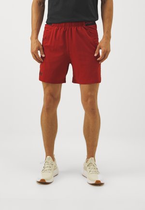 Mężczyzna noszący czerwone sportowe spodenki i beżowe buty do biegania stoi na jednolitym jasnym tle.