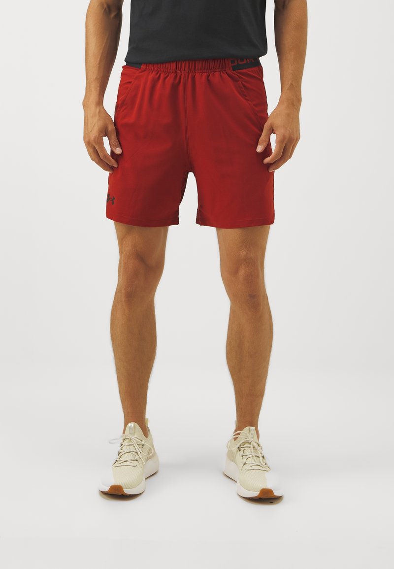 Homme portant un short de sport rouge et des chaussures de course beiges, debout devant un fond clair uni.