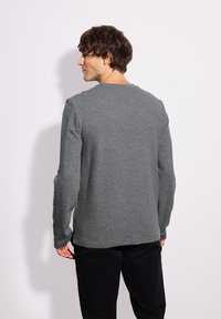 Maglietta grigia a maniche lunghe con finitura texture, scollo rotondo e cuciture a costine sui polsini e sull'orlo, abbinata a pantaloni scuri.