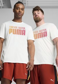 Balti trumpai rankovės marškiniai su spalvingu tekstu "AMŽINAI. GREIČIAU. PUMA". Abu modeliai dėvi raudonus šortus su Puma logotipu ir akcentais.