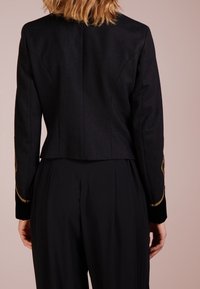 Černá krátká bunda s dlouhými rukávy, zdobená zlatou výšivkou na manžetách a s texturovaným povrchem, nošená s černými kalhotami s vysokým pasem.