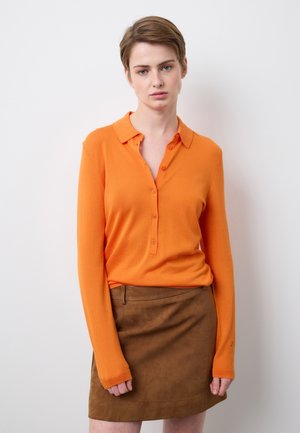 Mujer joven con cabello castaño corto, vistiendo una camisa naranja de manga larga abotonada y una falda de ante marrón, de pie frente a una pared blanca.