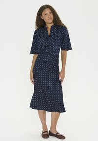 Robe bleu marine à manches courtes, col montant et pois blancs. Jupe évasée de longueur mi-genou, fabriquée dans un tissu lisse, assortie à des ballerines marron.