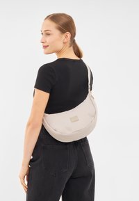 Borsa a tracolla in tessuto beige con forma arrotondata, dotata di un'etichetta frontale e di una tracolla regolabile, portata sulla spalla. T-shirt nera e jeans.