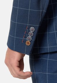 Puño de chaqueta de traje a cuadros azul oscuro con cuatro botones: tres grises con detalles en rojo y uno marrón claro. Costura en rojo a lo largo de la unión.