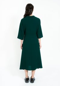 Tmavozelené šaty s dlhým rukávom a golierom, strednej dĺžky do polovice lýtok, s hladkou textúrou a minimalistickým dizajnom. Čierne topánky nosené pod nimi.