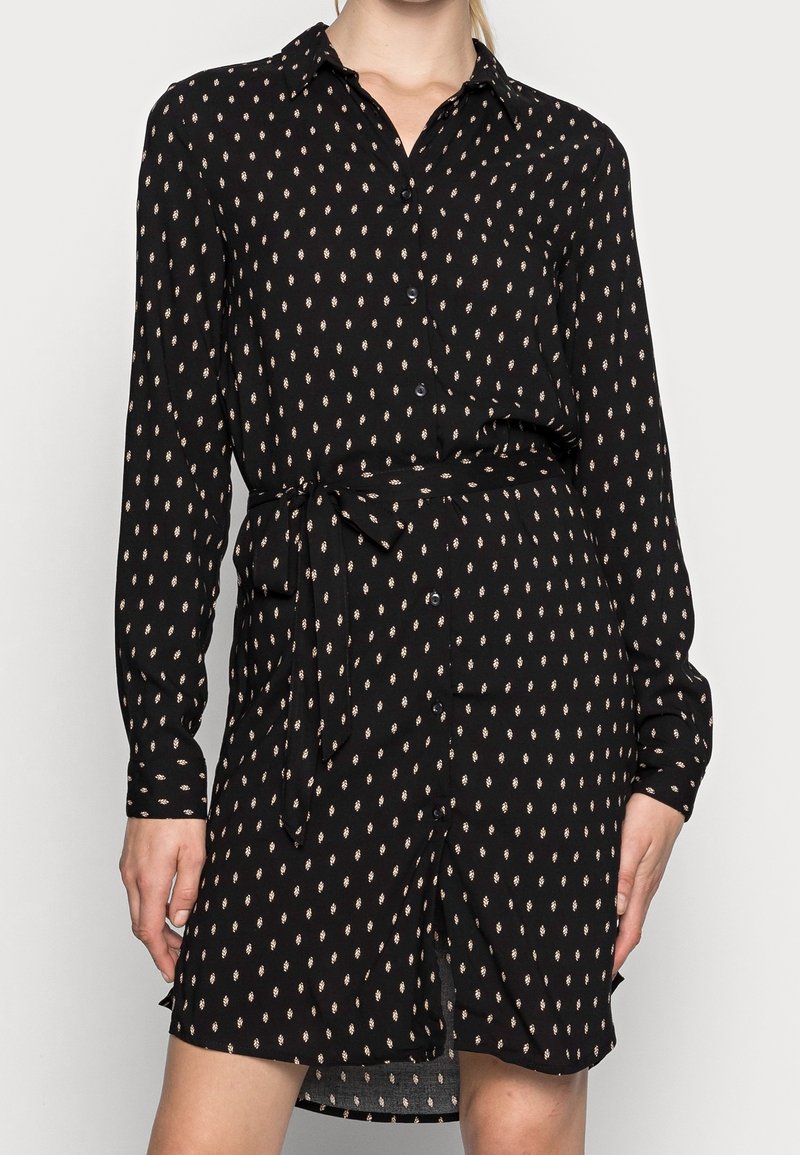 Femme portant une robe noire à manches longues boutonnée, avec de petits motifs floraux clairs et une ceinture à nouer à la taille.
