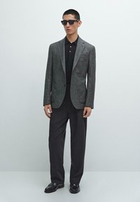 Gray herringbone blazer, black polo shirt, dark trousers, and black loafers. The blazer features a notched lapel and two front pockets.