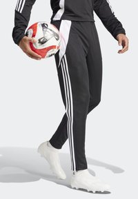 Svarta träningsbyxor med vita ränder, kombinerade med en svart långärmad topp och vita skor, håller en röd och vit fotboll.