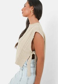 Pull en tricot beige sans manches avec un col montant, des motifs texturés et des liens sur les côtés. Assorti à un short en denim taille haute.