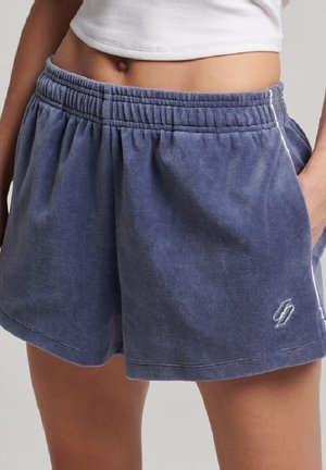 Personne portant un short en éponge à taille élastique bleu avec un petit logo blanc, debout avec une main dans la poche et un haut blanc court visible.