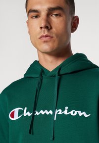 Groene hoodie van katoen, met een vooraanstaand logo in witte cursieve letters en een klein rood accent, voorzien van een capuchon met trekkoord en een gestructureerde stof.