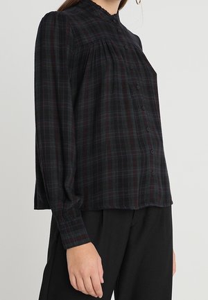 Kvinde iført en sort og mørk ternet langærmet knapbluse med flæset krave og sorte bukser, stående mod en ensfarvet baggrund.