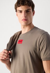 Tricou din bumbac khaki cu guler rotund și un patch logo roșu rectangular cu inscripția "HUGO" pe piept; textură netedă.