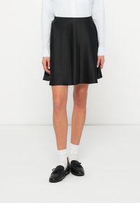 Crna satenska suknja s raširenim oblikom i glatkom teksture, u kombinaciji sa svijetloplavom prugastom košuljom i crnim cipelama preko bijelih čarapa.