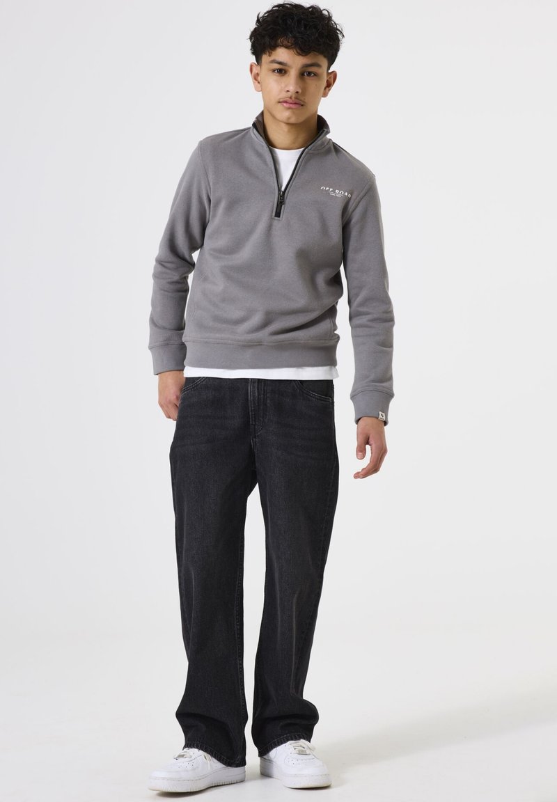 Chico adolescente de pie, vistiendo un suéter gris de manga corta con cremallera, camisa blanca debajo, jeans negros y zapatillas blancas, contra un fondo blanco liso.