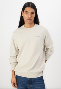 Felpa beige chiaro con collo a giro. Presenta polsini e orlo a coste. Logo discreto nell'angolo sinistro superiore. Indossata con jeans di denim scuri.