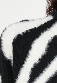 Pulover negru tricotat cu dungi groase albe zigzag, pufoase, pe umăr, cu păr creț și închis la culoare vizibil parțial pe lateral.