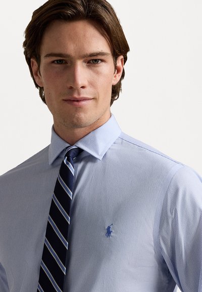 Chemise habillée rayée bleu clair avec un col pointu et une cravate rayée navy, ornée d'un petit logo brodé sur la poitrine gauche.